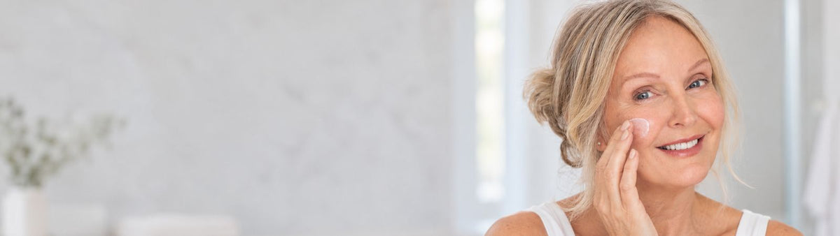 Mature woman gently applying moisturiser to her cheek in a softly lit bathroom.