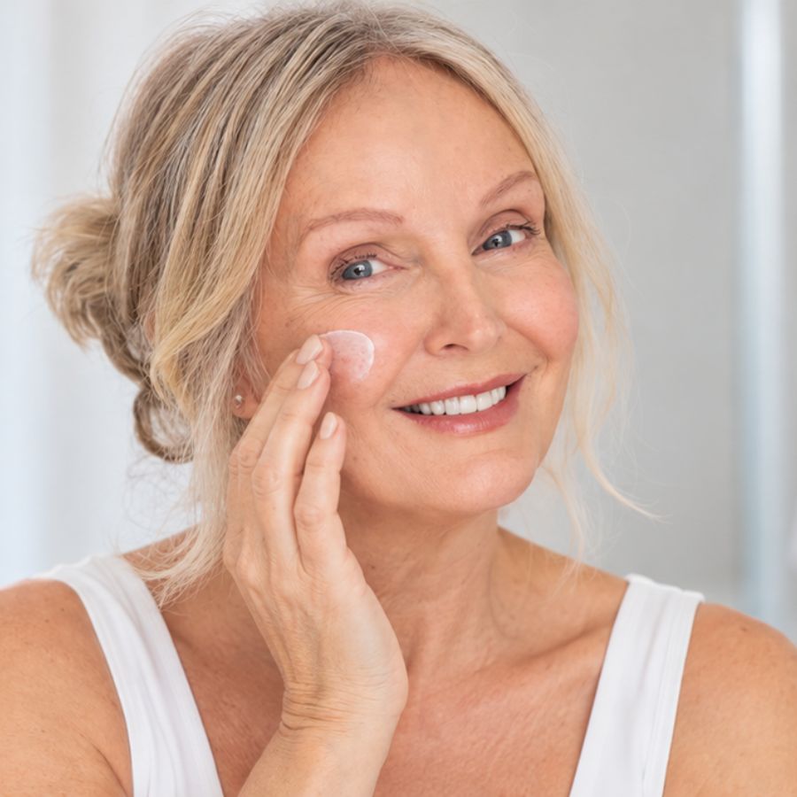 A smiling older woman with blonde hair in a bun applies a dollop of white face cream to her cheek. She is wearing a white tank top against a bright, soft-focus indoor background.