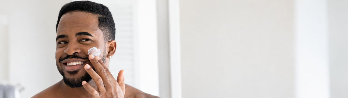 Man smiling while gently applying moisturiser to his cheek in a bright bathroom.